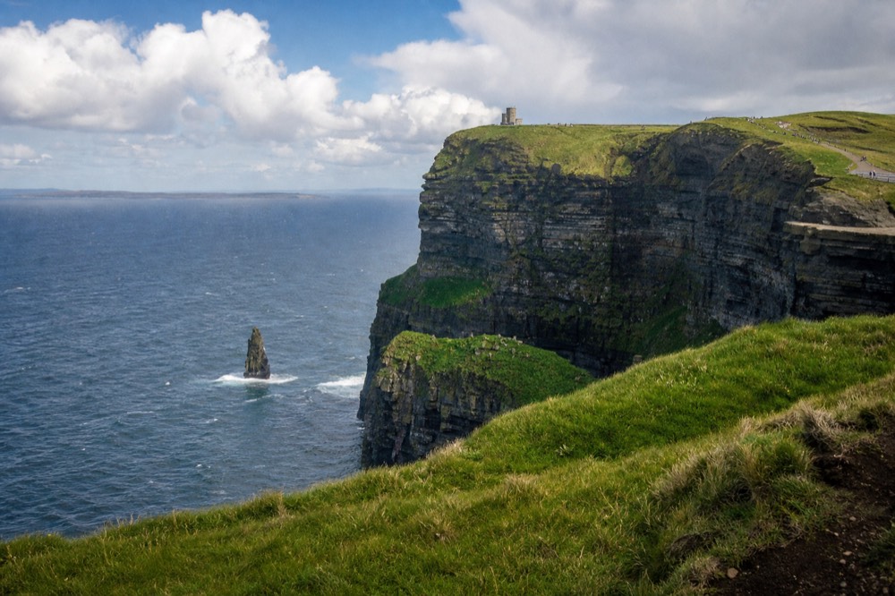 A wide view along the Cliffs of Moher on the Clare mainland, O'Brien's Tower on the high headland and the layered dark rock wall dropping to the Atlantic surf below.