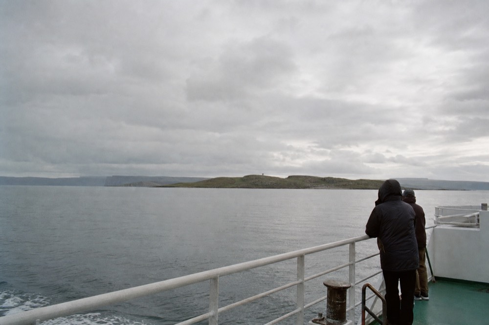 A small ferry crossing calm water between the Clare mainland and Inis Oírr, the green island ahead and the faint dark line of distant mainland cliffs on the horizon.