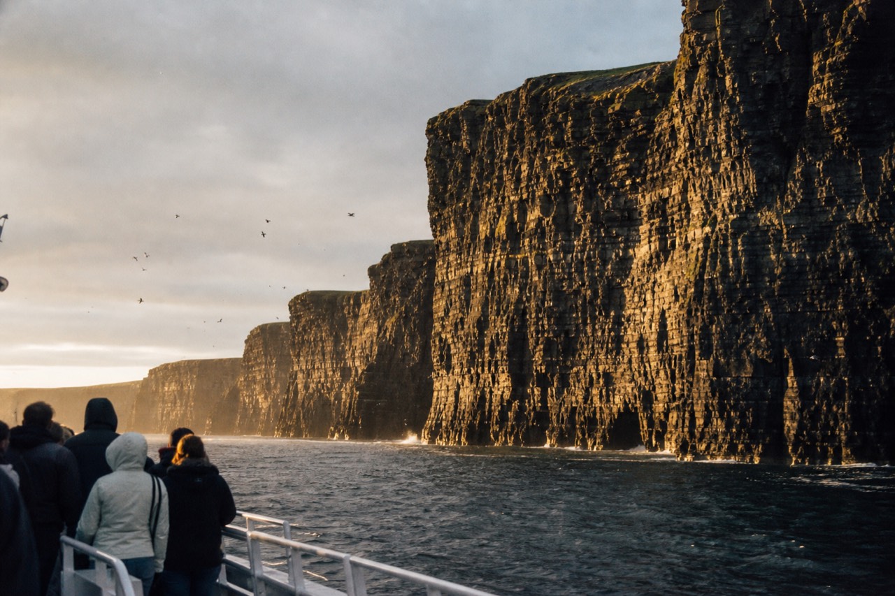 A small passenger ferry tracking the base of the Cliffs of Moher, the dark layered rock rising far above the deck and a scatter of seabirds over the swell.
