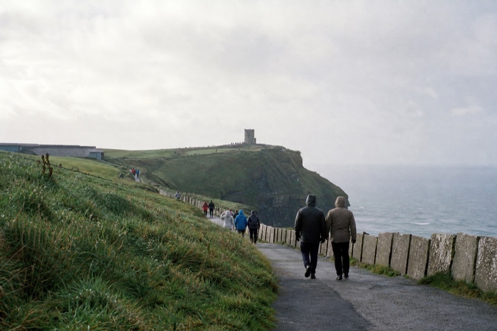 Visitors walking the clifftop path toward O'Brien's Tower at the Cliffs of Moher on a bright day, the visitor centre grassed into the hillside behind them.