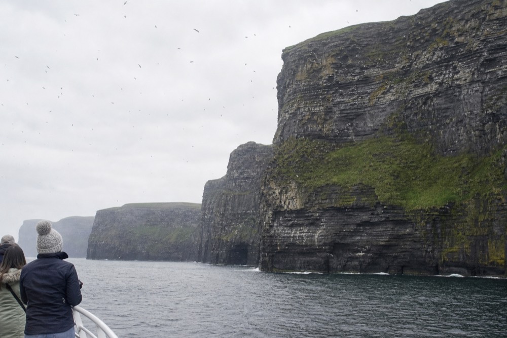 The Cliffs of Moher rising sheer from the Atlantic, viewed from a cruise boat at their base, the dark rock streaked with green and a haze of seabirds overhead.