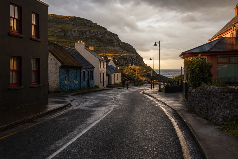 A quiet Doolin street at golden hour with painted cottages and a pub, the Burren hills rising behind and the sea a thin bright line at the end of the road.