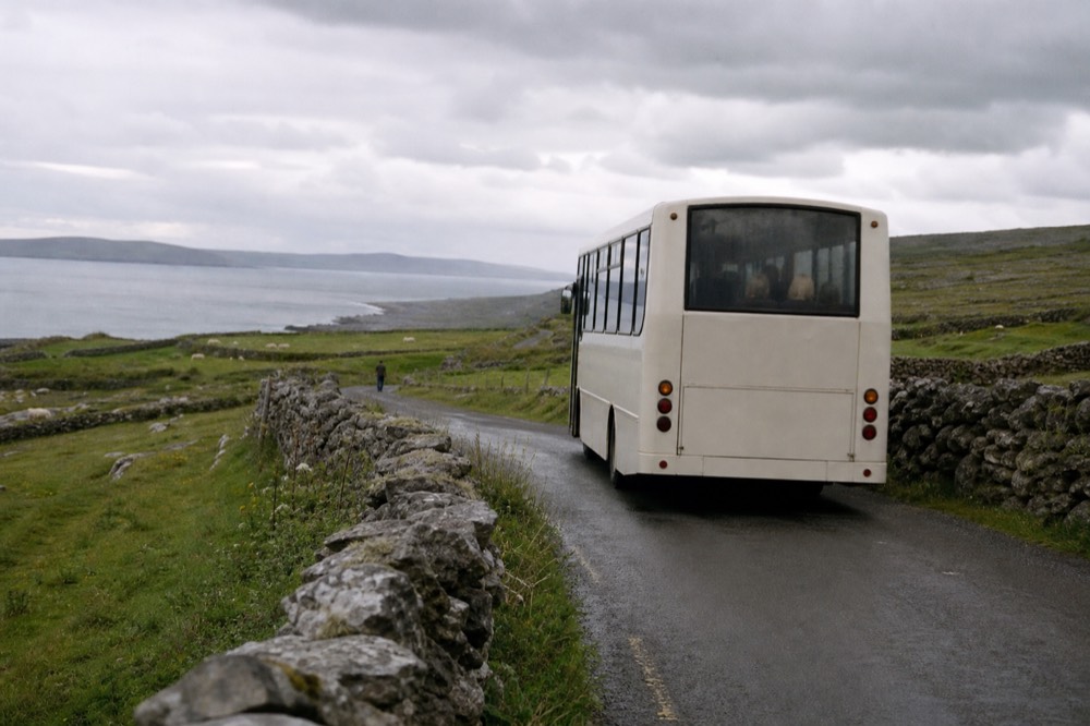 A regional bus on a narrow coastal road in County Clare, grey stone walls and green Burren fields on either side, the Atlantic showing between low hills.