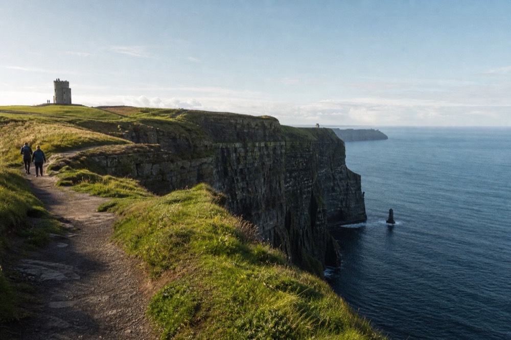 A quiet clifftop path at the Cliffs of Moher in early morning light, only a couple of walkers near the edge, O'Brien's Tower on the skyline above the empty sea.
