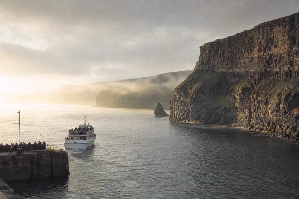 Early morning light on a calm Atlantic below the Cliffs of Moher in May, a small cruise boat leaving Doolin Pier with mist lifting off the green headland.