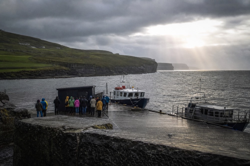The stone quay at Doolin Pier with two small tour boats moored, passengers in raincoats gathered near a ticket kiosk, green Clare hills behind and the grey Atlantic ahead.