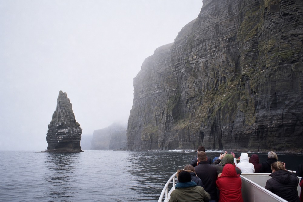 A cliff cruise boat close under the base of the Cliffs of Moher, passengers looking up at the towering rock face, a tall sea stack rising from the water nearby.