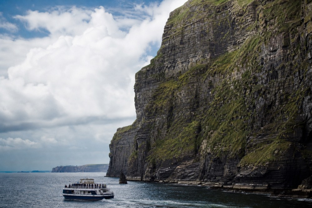 A cliff-cruise boat dwarfed at the base of the Cliffs of Moher, passengers along the rail looking up at the towering layered rock face streaked with green.