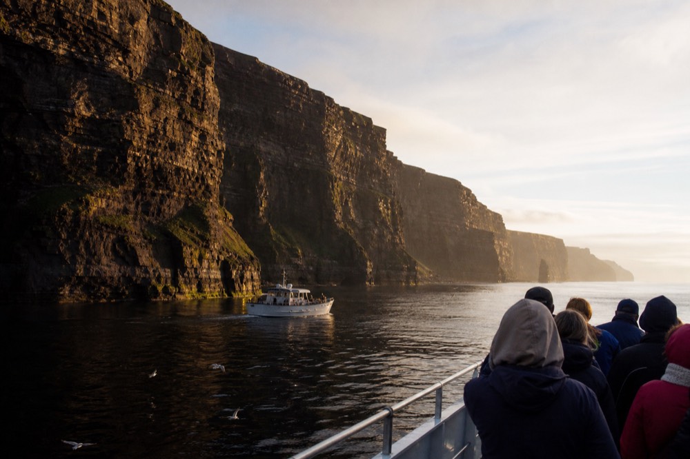 A calm early-summer morning at the Cliffs of Moher seen from the water, the sea almost flat, soft light on the layered rock and a few seabirds low over the surface.