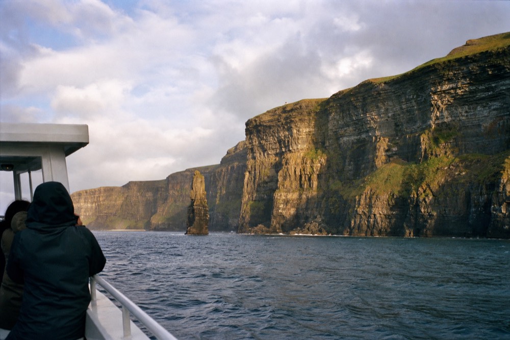 A late-spring morning on the water below the Cliffs of Moher, the boat steady on gentle swell, warm light across the full height of the layered rock wall.