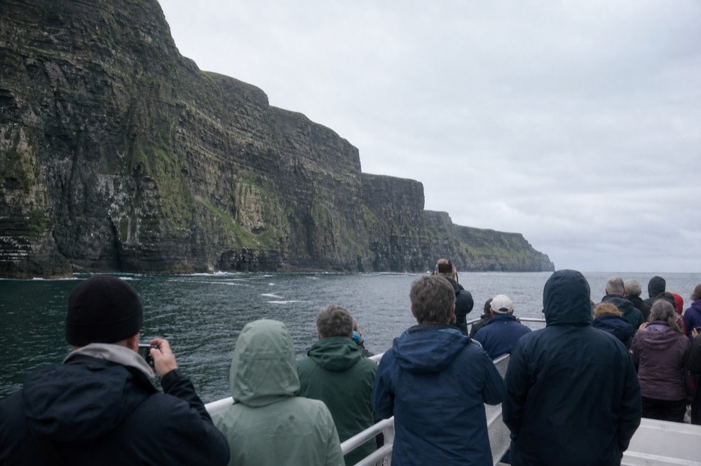 Passengers on the open deck of a cliff-cruise boat in soft June light, some photographing the towering cliffs, the sea calm and the sky bright behind the wall.