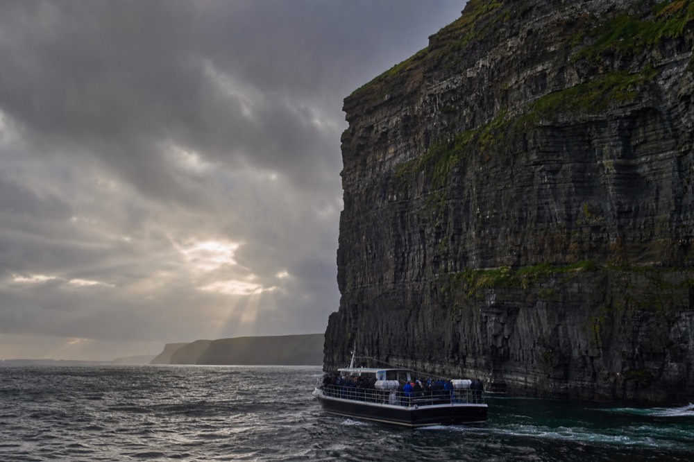 A cliff-cruise boat sitting low and close under the towering face of the Cliffs of Moher, passengers looking up at the layered rock wall rising hundreds of feet above them.