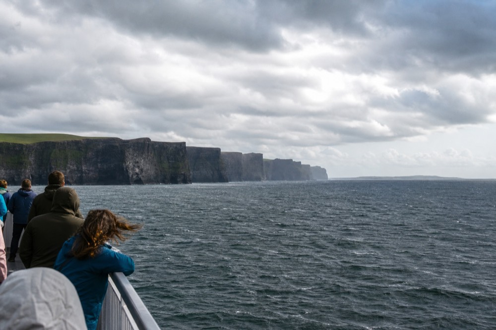 Passengers along the open deck of a returning boat looking back at the Cliffs of Moher receding behind them, the green Aran Islands low on the horizon in soft afternoon light.