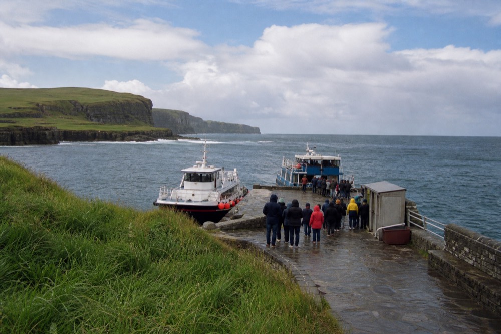 The stone quay at Doolin Pier with two small tour boats moored, passengers in raincoats waiting near a ticket kiosk, the grey Atlantic beyond.
