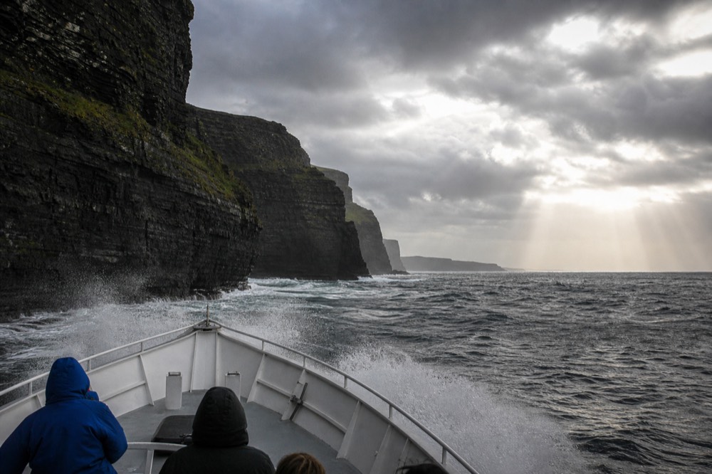 The bow of a cliff-cruise boat riding over a grey Atlantic swell at the base of the Cliffs of Moher, spray flying, the dark rock wall looming close on one side.