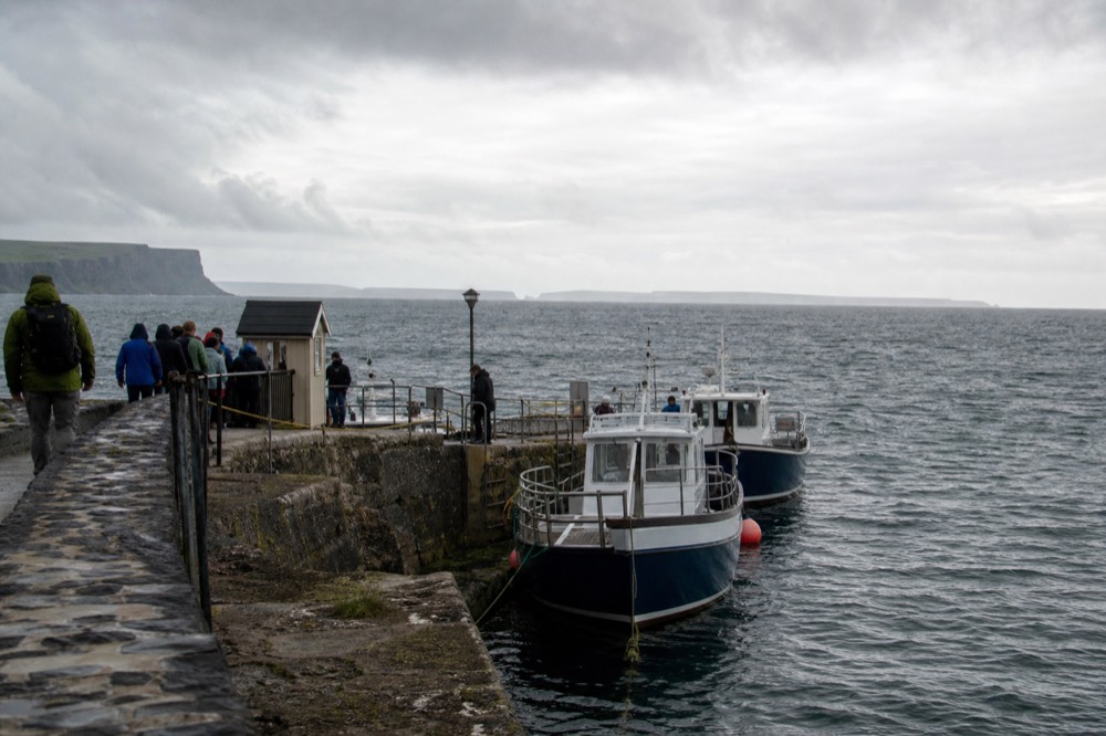The stone quay at Doolin Pier with two small tour boats moored, passengers in raincoats near a ticket kiosk, the grey Atlantic and the Aran Islands beyond.
