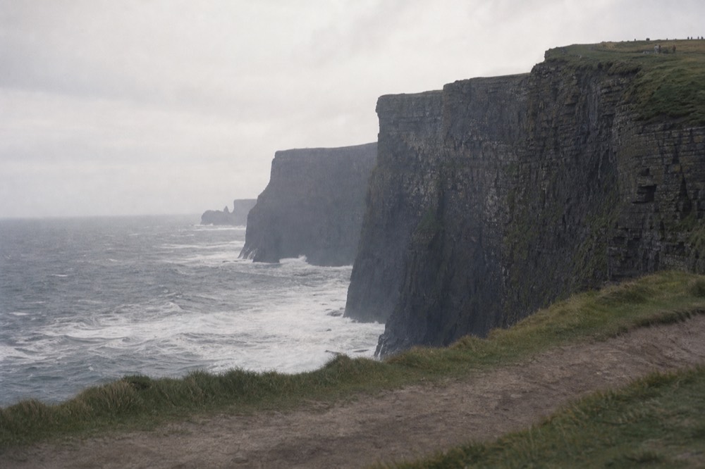 Choppy white-capped water churning at the base of the dark Cliffs of Moher, waves rebounding off the vertical rock face into a confused sea.