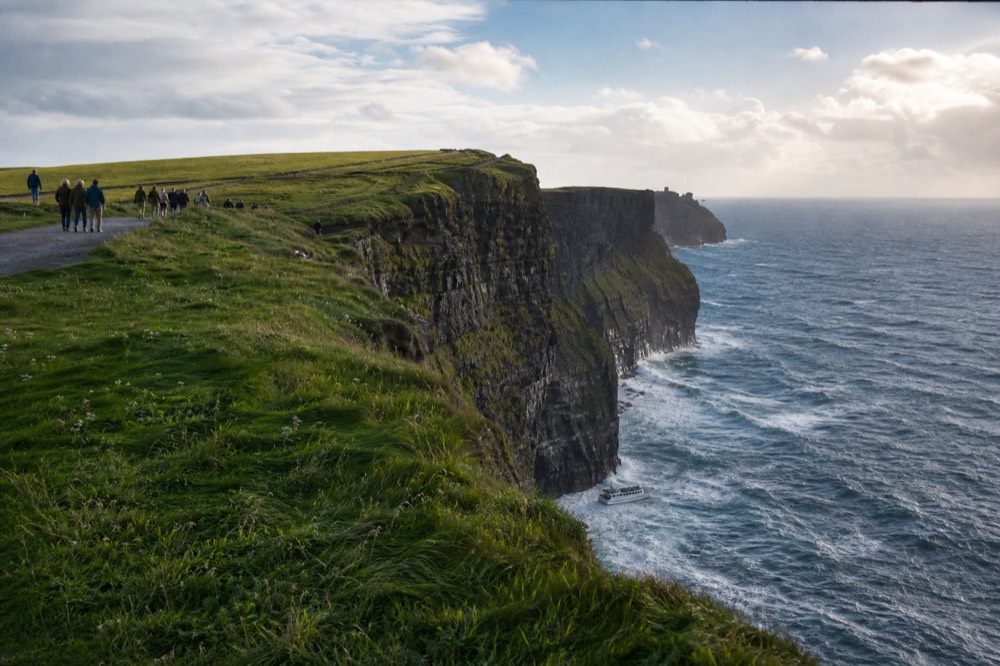 Split scene of the Cliffs of Moher: walkers small on the green clifftop path above, and a tour boat tiny at the base of the towering rock wall below them.