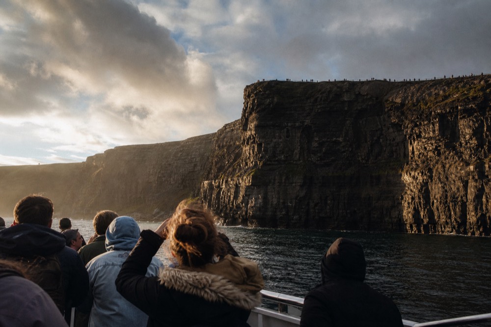 Passengers on a cliff-cruise boat looking up at the Cliffs of Moher while, far above on the clifftop, other visitors line the rail looking down at the water.
