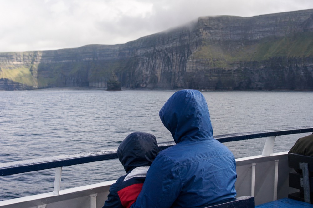 A parent and child in waterproofs sitting on the open deck of a tour boat, both looking calmly out at the flat sea and the cliff wall on a settled morning.