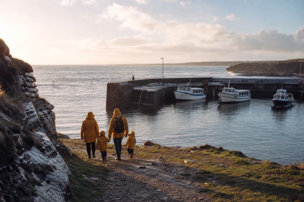 A family in matching raincoats walking down toward Doolin Pier with a small backpack, the harbour and moored tour boats ahead under a bright breezy sky.