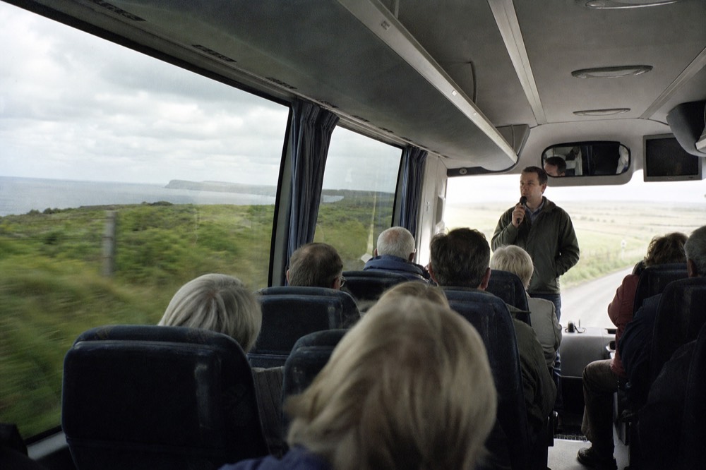 A guide standing at the front of a tour coach talking to seated passengers, green countryside blurring past the large windows on a bright morning.