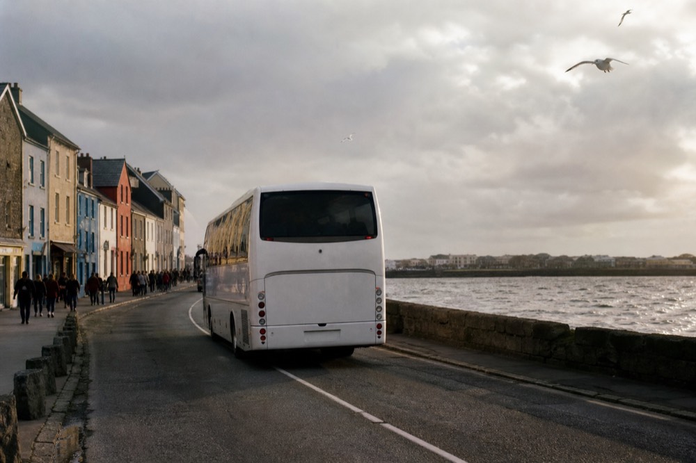 A tour coach pulling away from a busy Galway city street in the morning, colourful shopfronts and pedestrians behind it, the road heading south out of town.