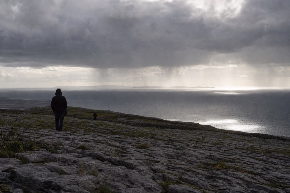 The pale limestone pavement of the Burren rolling toward the sea, cracked grey rock in the foreground and the faint outline of the Aran Islands on the horizon.