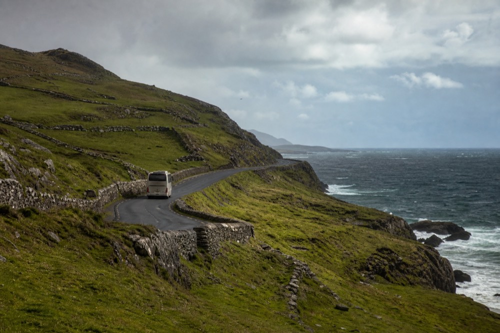 The coast road winding south from Galway with the Atlantic on the right and green stone-walled fields climbing to the left, a tour coach small in the distance.