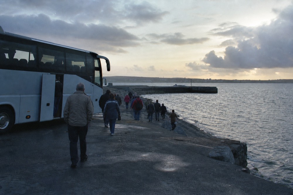 A returning tour coach parked at the Galway waterfront in the evening, passengers stepping down against a soft dusk sky over the bay.