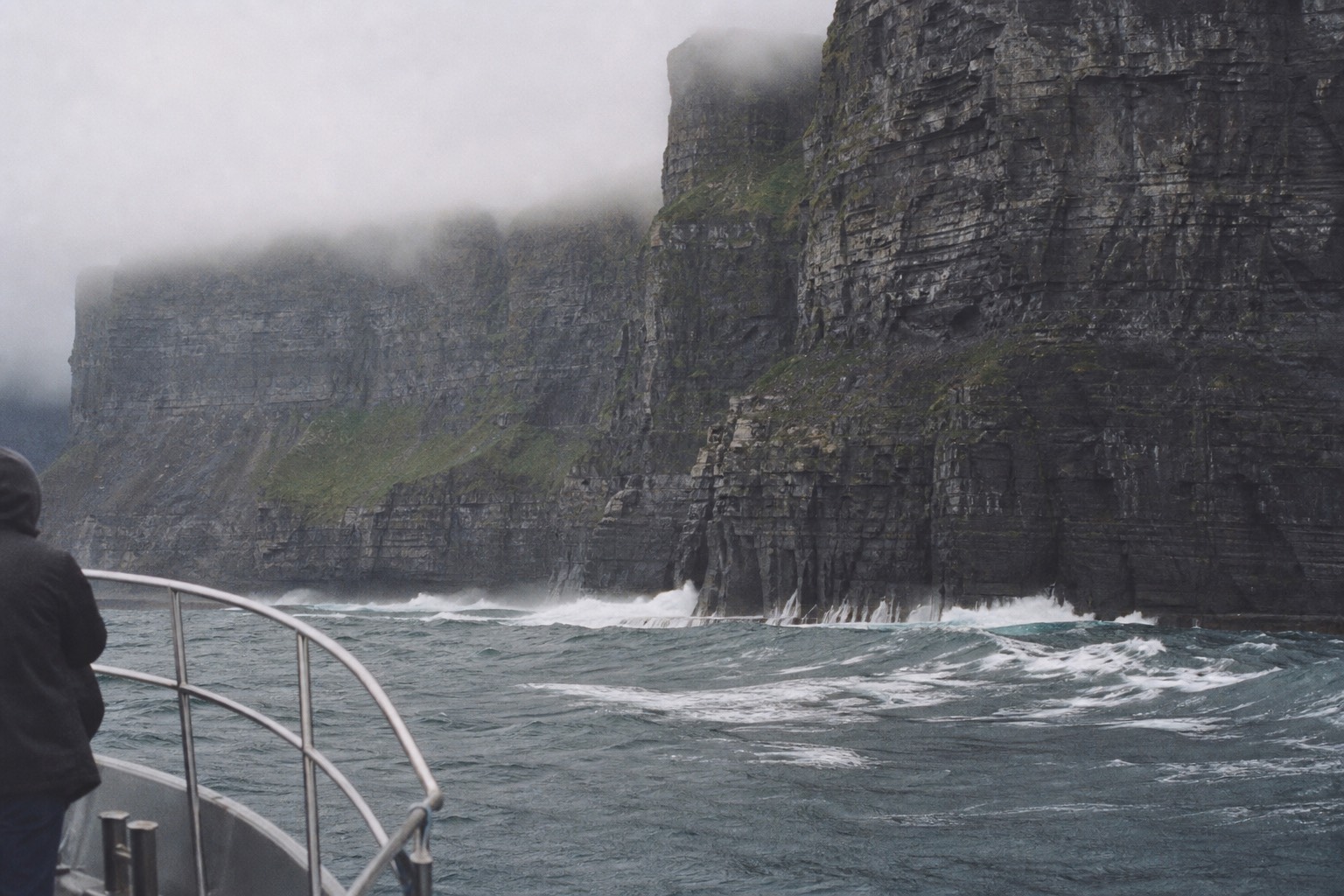 The Cliffs of Moher rising sheer from the Atlantic seen from sea level off a small boat, the dark wet rock face towering into low cloud, heavy ocean swell breaking white at the base under moody silver-grey Irish light